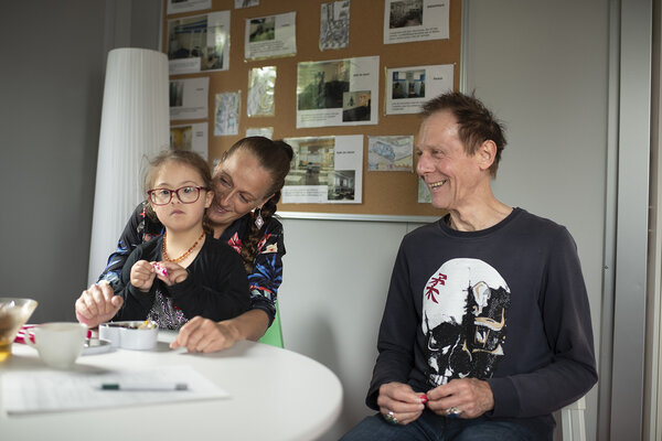 Scène d'échanges souriants entre un bénévole, une mère et sa petite fille, avant un parloir.